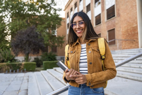 Young woman smiling, wearing glasses and a backpack, holding a book, and standing on university stairs, representing education, learning, and future success
