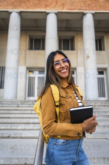 Young woman smiling, wearing glasses and a backpack, standing with a notebook in front of a university building's classical architecture, embracing education and student life