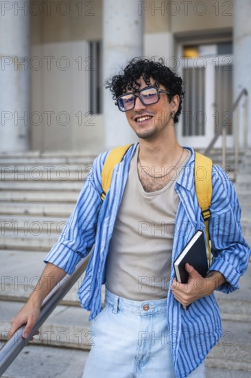 Young man with a backpack and a notebook, smiling while walking up the modern building stairs, representing education, collegiate life, and future opportunities