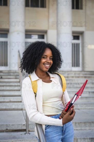 African american woman smiling while holding a notebook, wearing a backpack and standing in front of a university building with classical columns and stairs