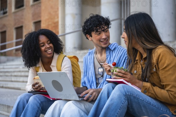 Group of multi ethnic university students sitting on outdoor steps, sharing a light meal, laughing, and interacting during a social break while using a laptop