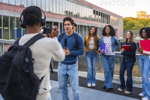 Student journalist filming and interviewing a young male student while a diverse group of female students watches, capturing a moment of campus media production