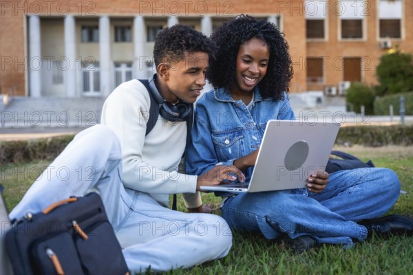 Young black students sitting on green grass, working on a laptop, discussing school project or online course content at campus, representing education, learning, and modern study