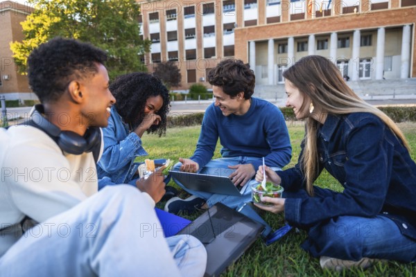 Group of diverse university students enjoying a refreshing outdoor lunch together on a grassy campus lawn, laughing and using a laptop with books nearby