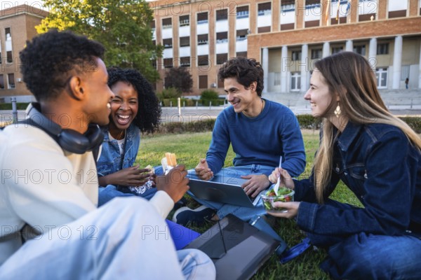 Group of happy young students smiling and laughing, sitting on grass having lunch and working with a laptop in front of a university building on a sunny day
