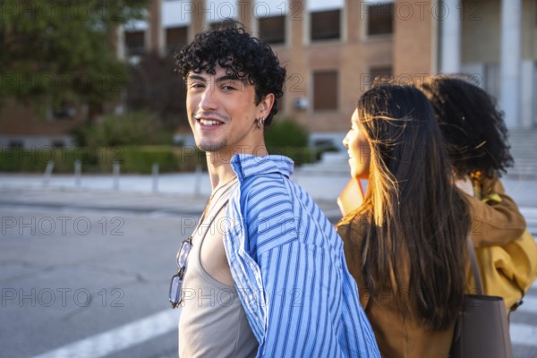 Young man with curly hair smiling at camera while walking with two female classmates on a sunlit university campus pavement, casual friends enjoying city college life