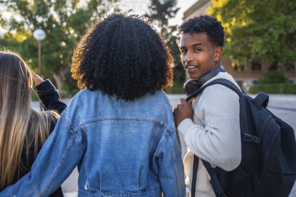Three diverse young university students walking together on campus, the smiling man looking back at the camera while wearing headphones and a backpack