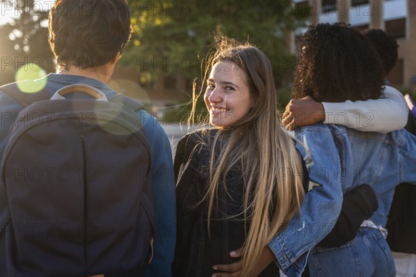 Group of young diverse university students walking arm in arm on campus, one woman turning to camera smiling during golden hour, celebrating friendship and education