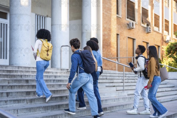 Group of multi ethnic students with backpacks and books walking up the steps of a university building, representing modern education and friendship on campus