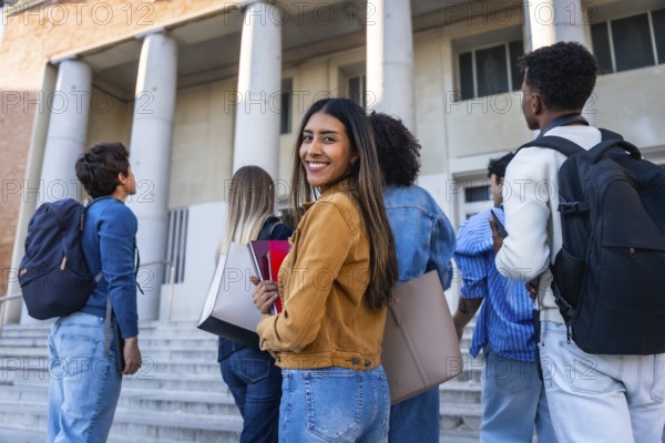 Group of young diverse students walking up steps towards a university building, carrying backpacks and books, with one smiling woman looking back at the camera