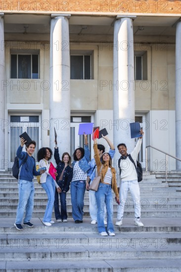 Diverse group of happy college students standing on the outdoor stairs of a university building, raising books and documents in the air, celebrating academic achievement and education