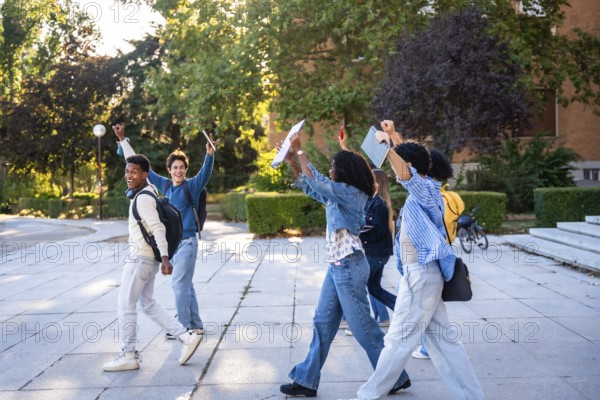 Diverse group of young university students walking on campus after classes, happily raising arms in celebration of academic achievement and friendship, enjoying a bright future