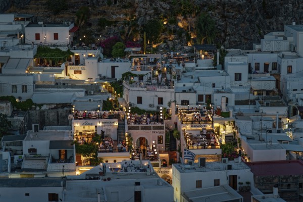 View of the illuminated roof terraces and restaurants of the town of Lindos and white houses that radiate a cosy atmosphere, Acropolis, Lindos, Rhodes, Island, Greece