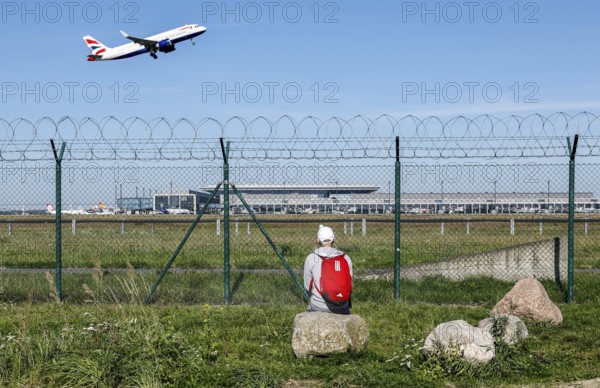 A man sits on a stone and watches the take-off of a British Airways plane through a barbed wire fence at BER Airport, Berlin Schönefeld, 28/09/2025, Schönefeld, Brandenburg, Germany