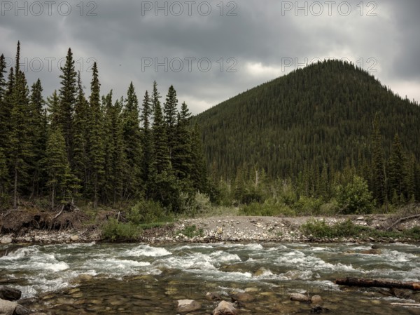 A rugged river flows through a forest with tall mountains under a dramatic sky within Bragg Creek Provincial Park, in Alberta, Canada, North America