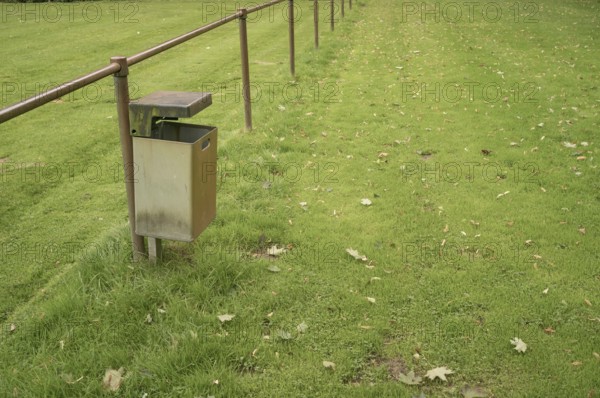 Borken, NRW, Germany, Empty metal garbage bin standing next to a railing in a public park with scattered autumn leaves