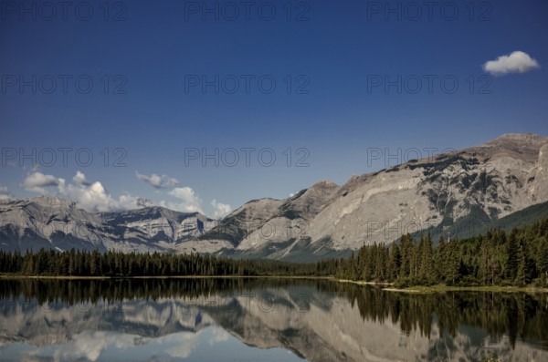 Reflections of rugged mountains of the Canadian Rocky Mountains in a calm lake framed by dense forest near Canmore, Alberta, Canada, North America