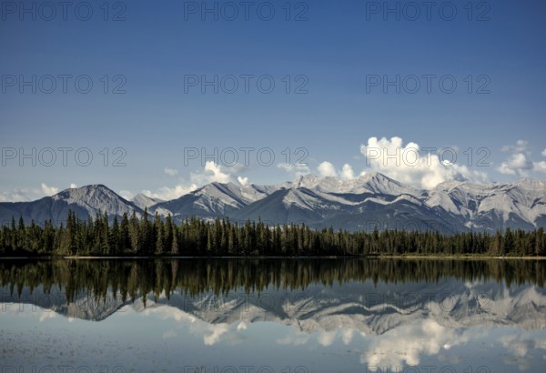 Tranquil lake mirroring mountains of the Canadian Rocky Mountains and sky, with pine forest and scattered clouds near Canmore, Alberta, Canada, North America