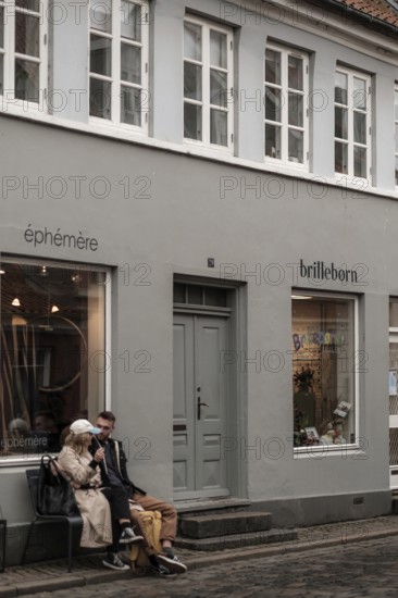 A couple sits on a bench outside a gray storefront having a conversation in Aarhus, Denmark, Northern Europe