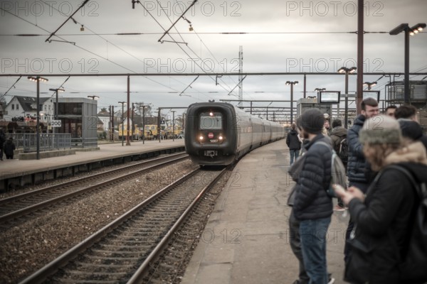 Commuters waiting at a platform as a train approaches, set against an overcast sky in Copenhagen, Denmark