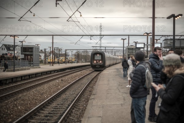Commuters waiting on a platform as a train approaches at an overcast train station in Copenhagen, Denmark