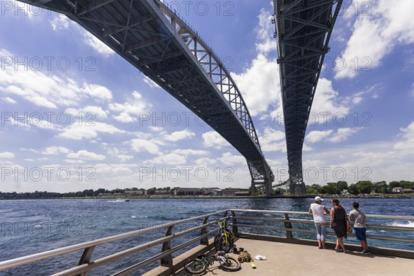 People standing on a riverside under the Blue Water Bridge, connecting Canada and the United States, with bikes nearby along the St. Clair River leading to Lake Huron in Sarnia, Ontario, Canada, North America