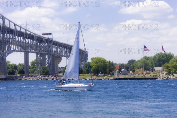 Sailboat gliding near the Blue Water Bridge, connecting Canada and the United States, with flags under a bright blue sky along the St. Clair River leading to Lake Huron in Sarnia, Ontario, Canada, North America