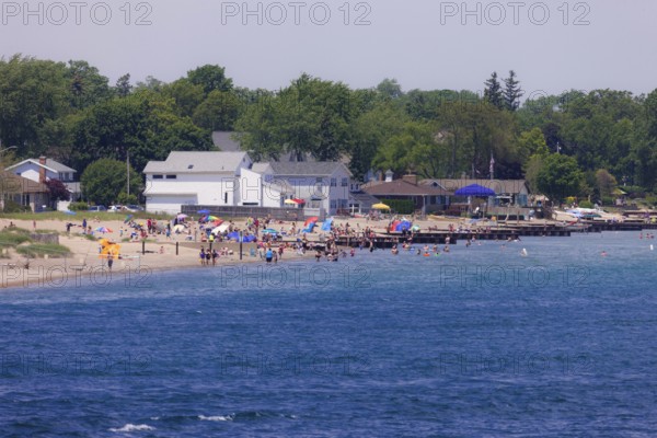 A busy beach scene with people enjoying the sun, umbrellas, and nearby houses along the St. Clair River leading to Lake Huron in Sarnia, Ontario, Canada, North America