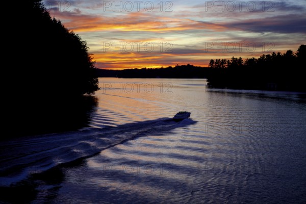 A boat moves across a lake during twilight, leaving ripples under a colorful sunset in Muskoka, Ontario, Canada, North America