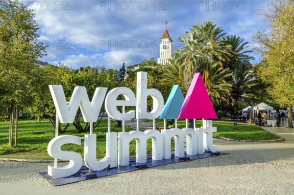 Nov 13, 2024 - Lisbon, Portugal: Large sign displaying Web Summit logo in lively park filled with greenery, historic tower in background. Major annual technology conference internet technology, emerging technology, marketing, AI solution, networking