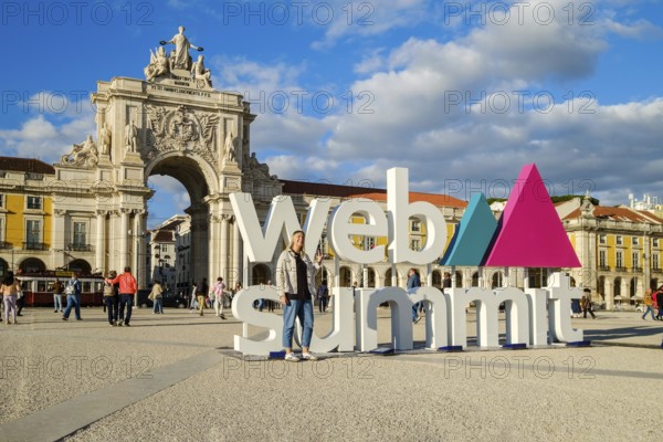 Nov 13, 2024 - Lisbon, Portugal: A female adult posing for a photo in front of Web Summit sign installation at Commerce square and Rua Augusta Arch. People walking, international technology conference, discussions, networking, technology, innovation