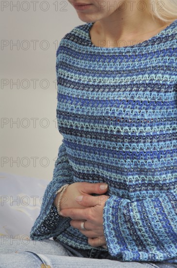 Young woman suffering, A woman in a blue striped sweater sitting indoors, clutching her hands over her stomach