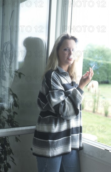 A woman with a striped shirt stands by a window, holding a cigarette, with natural light illuminating her