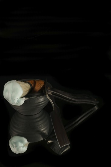 Shaving brush with cream and straight razor reflecting on a black background