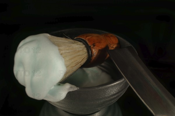 Shaving brush with cream and straight razor resting in a bowl on black background