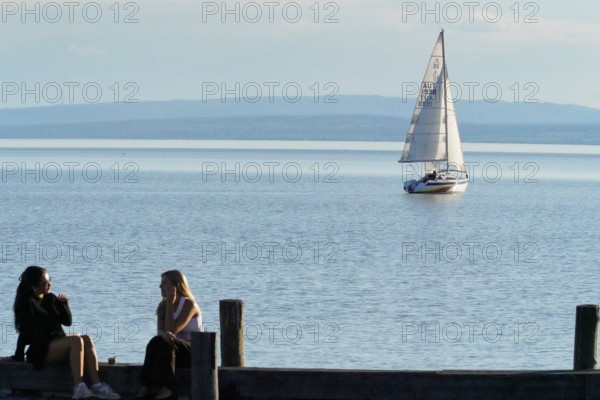 Lake Neusiedl, Burgenland, Austria, Europe, Two woman sit on the jetty while a sailing boat passes by on the calm lake, Sailing boats on Lake Neusiedl