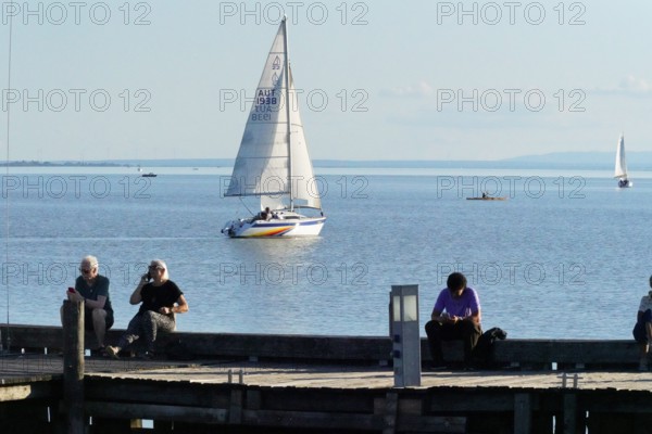 Lake Neusiedl, Burgenland, Austria, Europe, People sitting at the jetty and watching a sailing boat in the calm water under a blue sky, Sailing boats on Lake Neusiedl