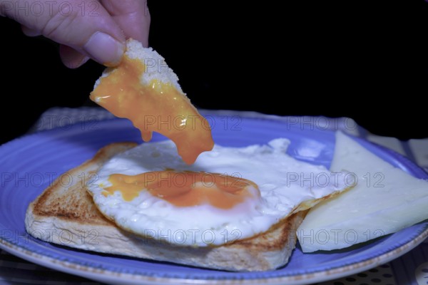Close-up of a hand dipping bread into a runny egg yolk, served on toast with cheese, creating a delicious and simple breakfast