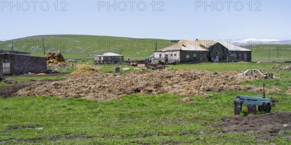 A farm with old buildings and agricultural machinery on a green pasture, cow dung in front of it, Berdashen, Lake Arpi, Shirak province, Armenia