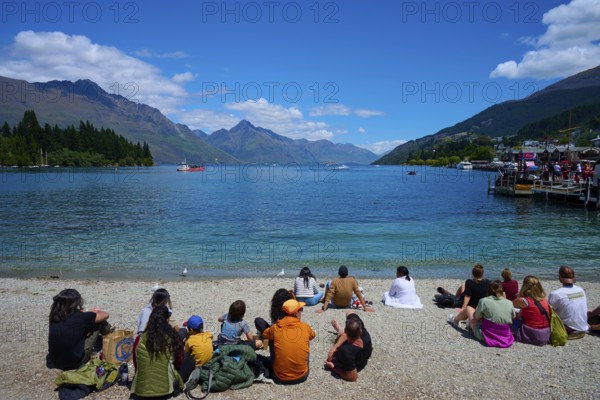 Groups of people sitting on the beach enjoying the view of the lake and mountains, Queenstown, Queenstown-Lakes District, Otago, South Island, New Zealand