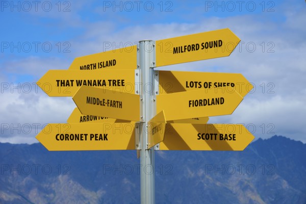 Yellow signpost with several signs and a mountain ridge in the background against a blue sky, Queenstown, Queenstown-Lakes District, Otago, South Island, New Zealand