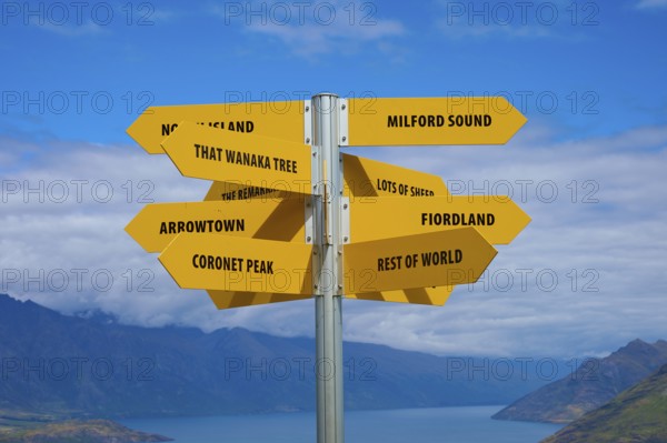 Yellow signpost with several signs in front of a mountain backdrop under a cloudy sky, Queenstown, Queenstown-Lakes District, Otago, South Island, New Zealand