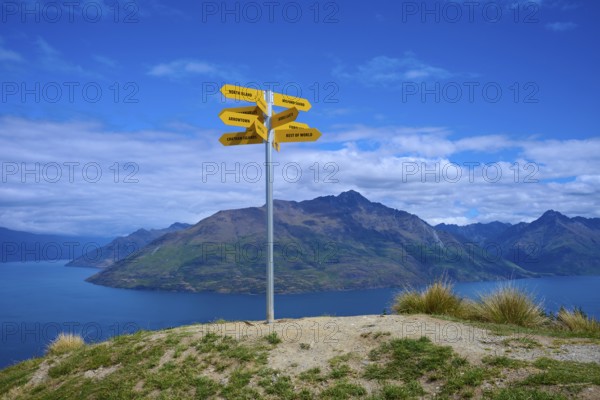 Yellow signposts on a hill in front of a vast mountain landscape under a blue sky, Queenstown, Queenstown-Lakes District, Otago, South Island, New Zealand