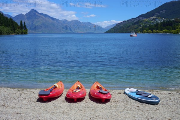 Red kayaks and paddleboard on the beach in front of a calm lake with mountain views, Queenstown, Queenstown-Lakes District, Otago, South Island, New Zealand