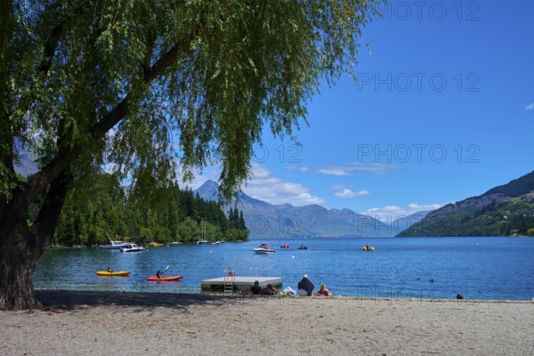 Sunny day at the lake with calm water, people relaxing on the shore and boats floating on the water, Queenstown, Queenstown-Lakes District, Otago, South Island, New Zealand