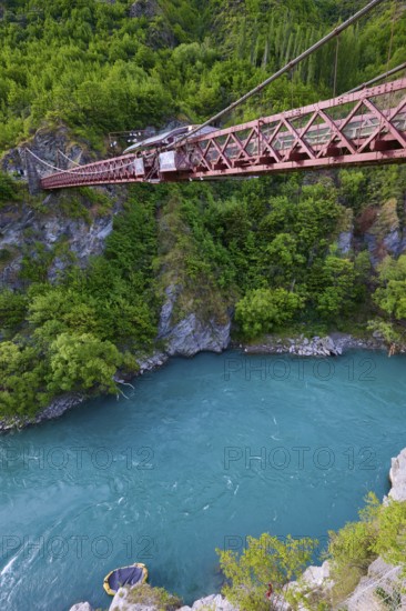 Red suspension bridge over river with steep wooded cliffs and calm waters, Kawarau Gorge Suspension Bridge, Kawarau River, Queenstown, Otago, South Island, New Zealand