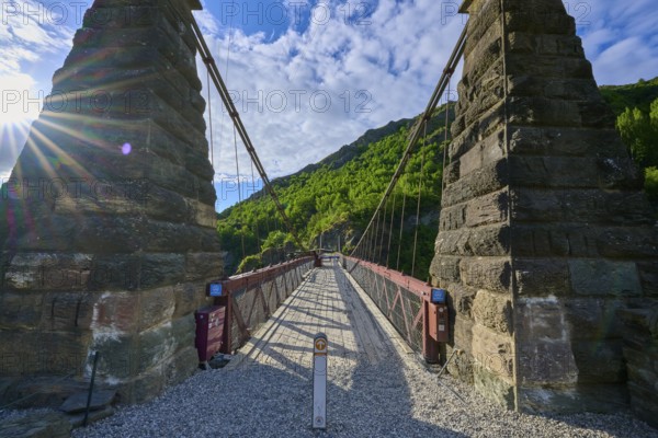 Old bridge with massive stone pillars surrounded by lush greenery under a bright blue sky, Kawarau Gorge Suspension Bridge, Kawarau River, Queenstown, Otago, South Island, New Zealand