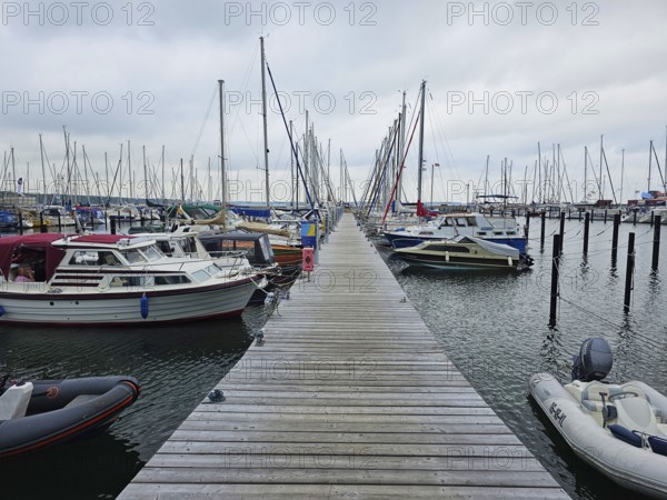 A wooden walkway leads through a harbour, lined with boats and masts under a cloudy sky, Laboe, Kiel Fjord, Schleswig-Holstein, Germany