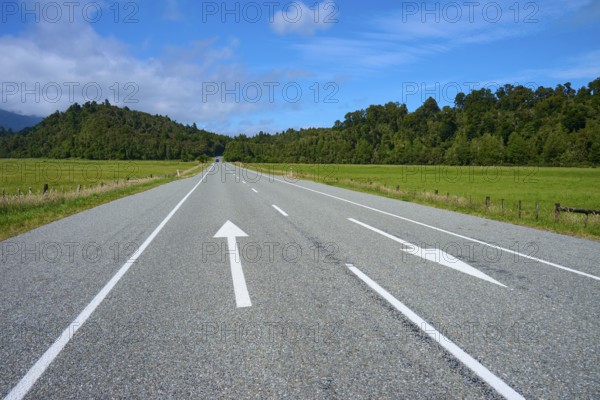 Wide road with arrows leads through green landscape under blue sky, West Coast, South Island, New Zealand