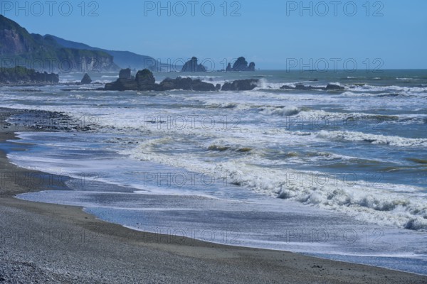 Dynamic waves roll onto a rocky beach under a bright blue sky, West Coast, South Island, New Zealand